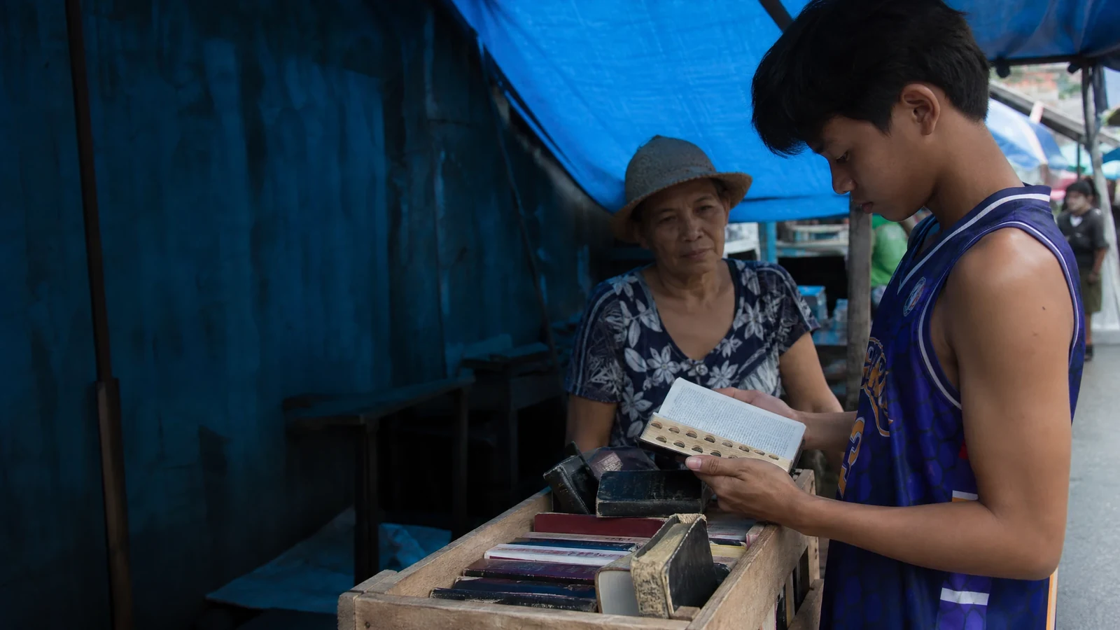 Un adolescente lee una pequeña Biblia usada en un puesto cubierto de un mercado mientras una mujer mayor lo observa