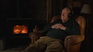 An old man with a white moustache listens on headphones in a leather armchair, eyes shut, by the glow of a wood stove.